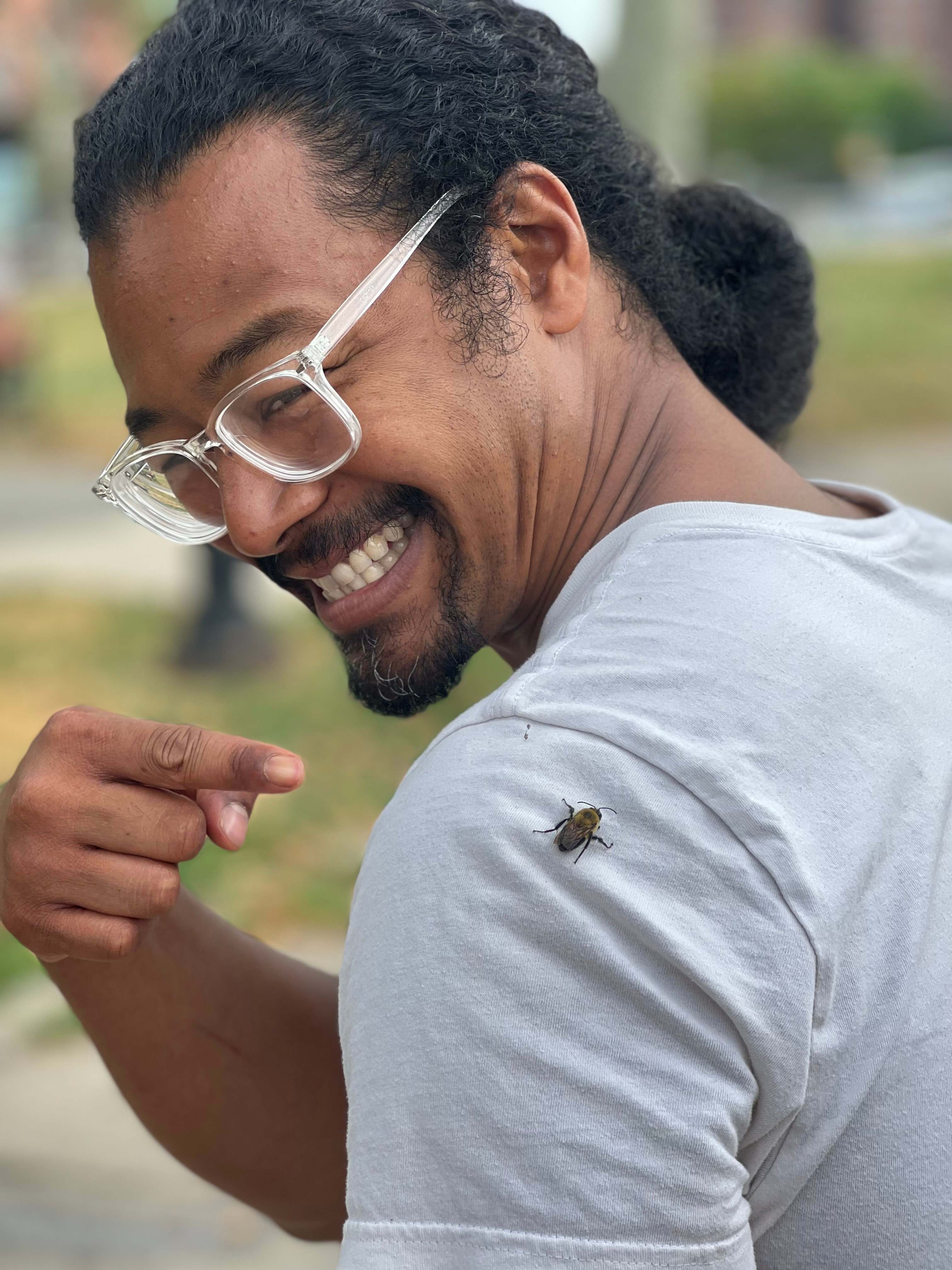 a photo of Julien Mellon in a white t-shirt and clear-framed glasses smiling while pointing at a bee that has landed on his shoulder